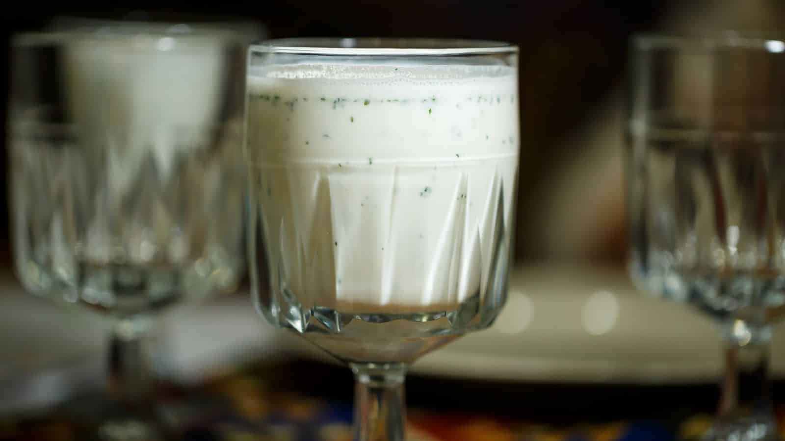 A clear, textured glass filled with a creamy, white liquid that has visible specks of green. Two similar glasses are blurred in the background. The setting appears to be a dining table.