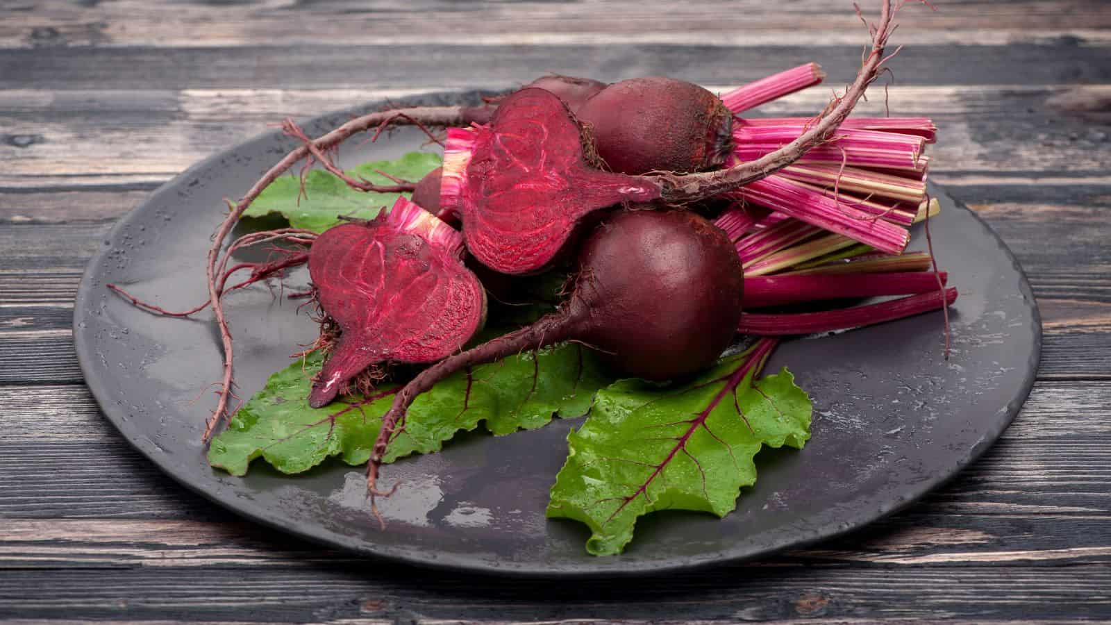 A dark plate holds four beetroots with leaves, placed on a wooden surface. Two whole beets and two halved beets show their deep red color, with pink stems and green leaves visible.