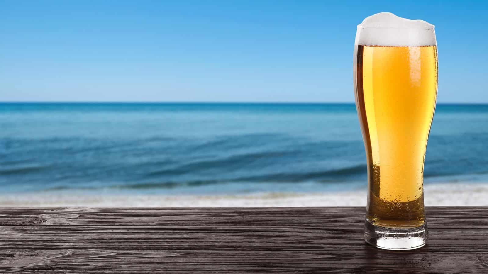 A tall glass of beer with a frothy head sits on a wooden table. In the background, a beach with gentle waves under a blue sky is visible.