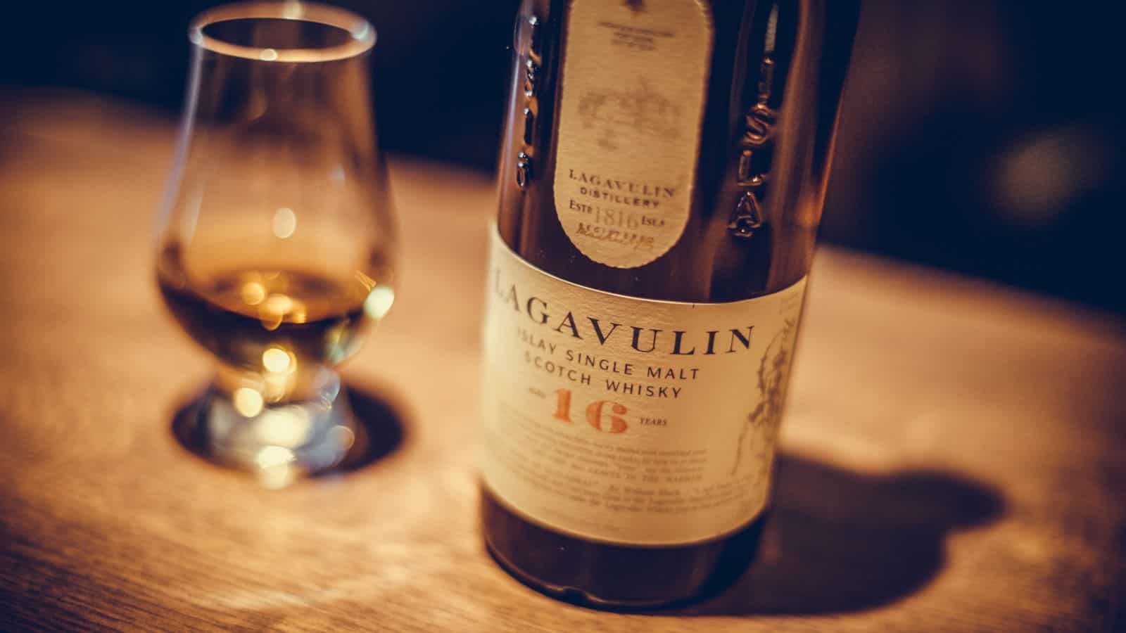 A bottle of Lagavulin 16-year-old Islay single malt Scotch whisky is displayed prominently with a partially filled whisky glass beside it on a wooden surface. The bottle’s label is clearly visible.