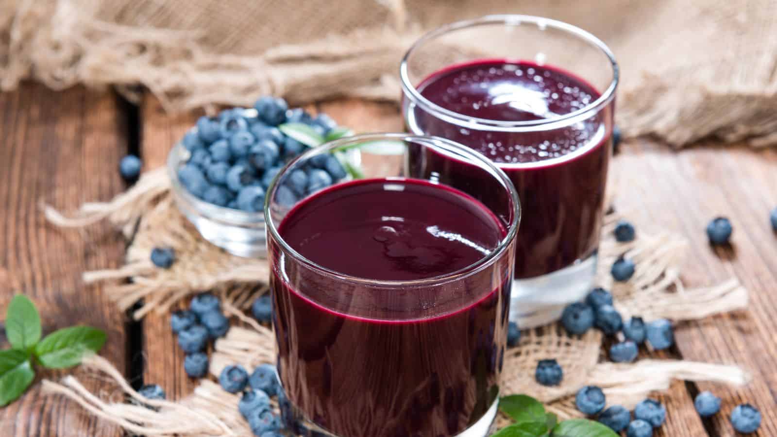 Two glasses of dark purple blueberry juice are placed on a wooden surface. A small bowl of fresh blueberries is nearby, with some scattered around. Burlap fabric is used as a rustic table setting. Green leaves are visible at the edges.