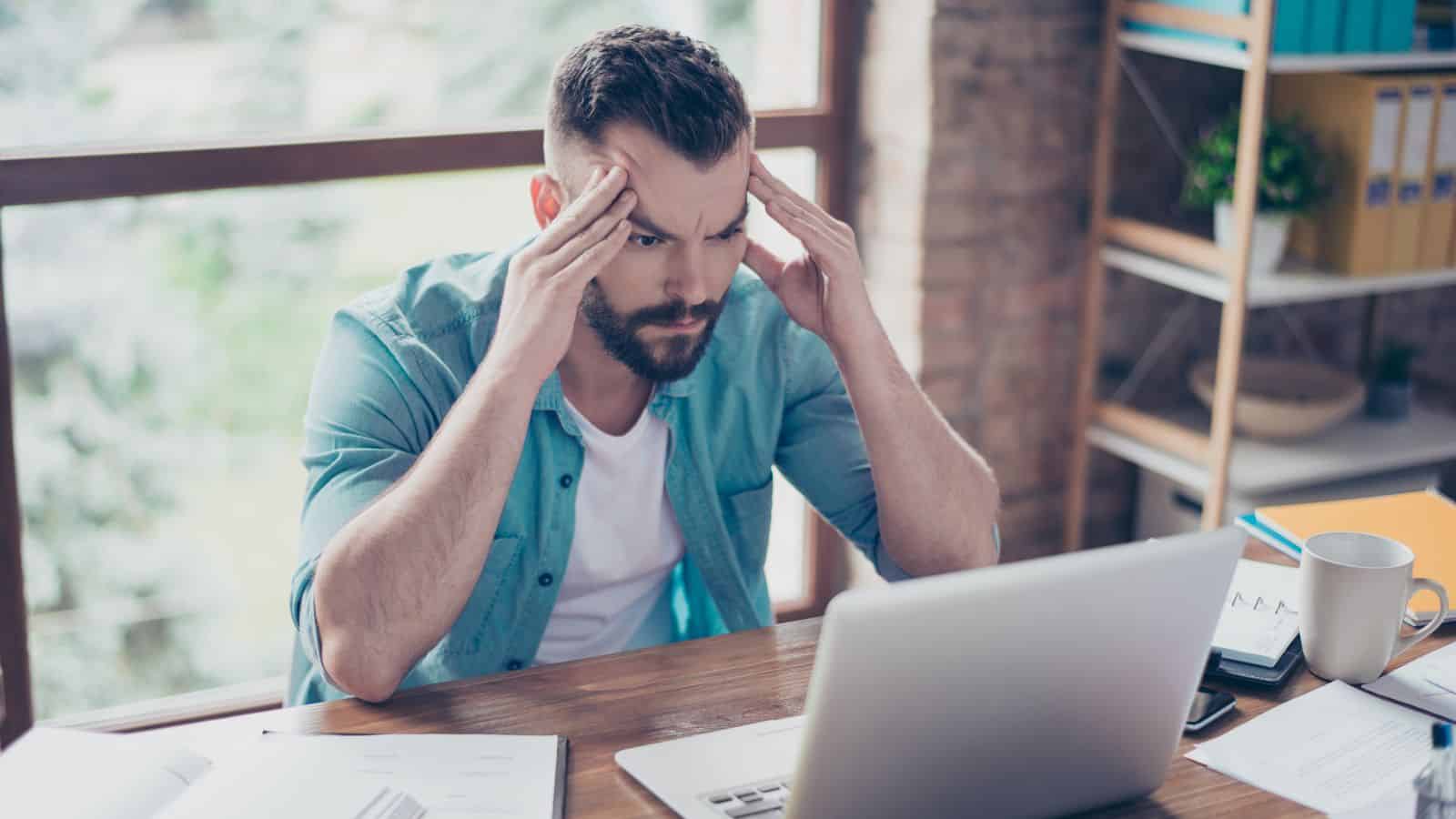 A bearded man in a teal shirt sits at a wooden table, looking stressed while holding his head with both hands, staring at a laptop. Papers, a smartphone, and a mug are on the table in a room with large windows.