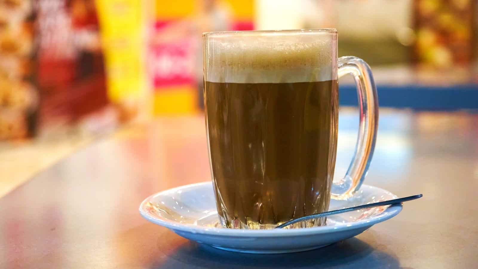 A tall glass mug of frothy coffee sits on a white saucer with a metal spoon beside it. The background is blurred with warm colors and shapes, suggesting an indoor caf&eacute; setting.