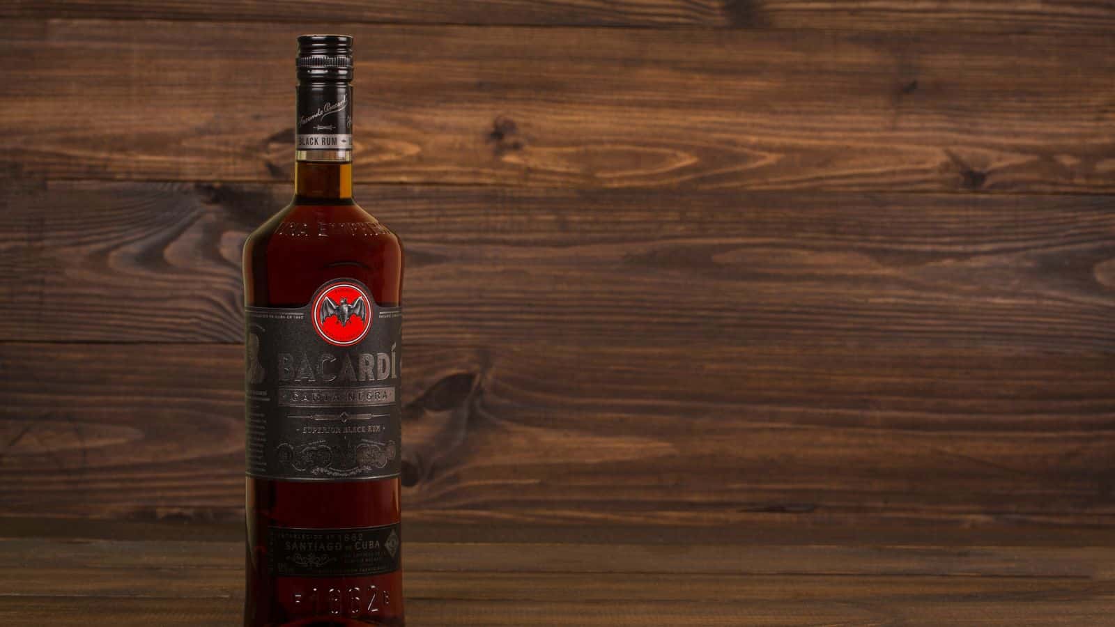 A bottle of Bacardi Carta Negra rum stands on a wooden surface against a wood-paneled background. The label is dark with silver lettering and a red bat logo near the top. The bottle has a black and silver cap.
