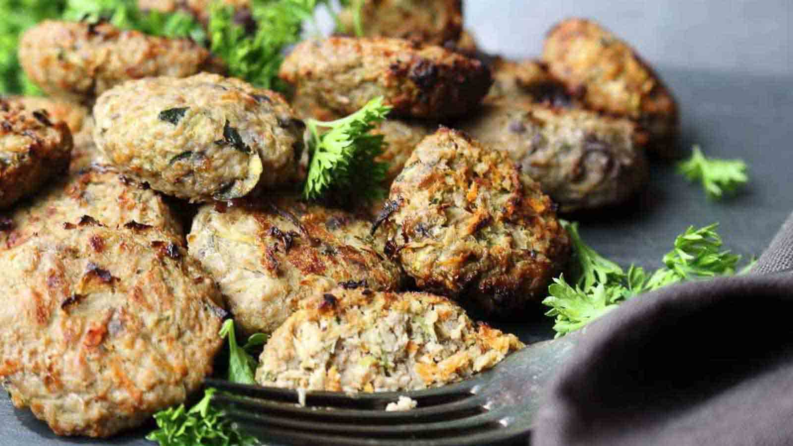 A close-up of several grilled meat patties with visible herbs, placed on a dark surface. Fresh parsley is scattered around for garnish. A metal fork is partially visible at the right edge of the image.