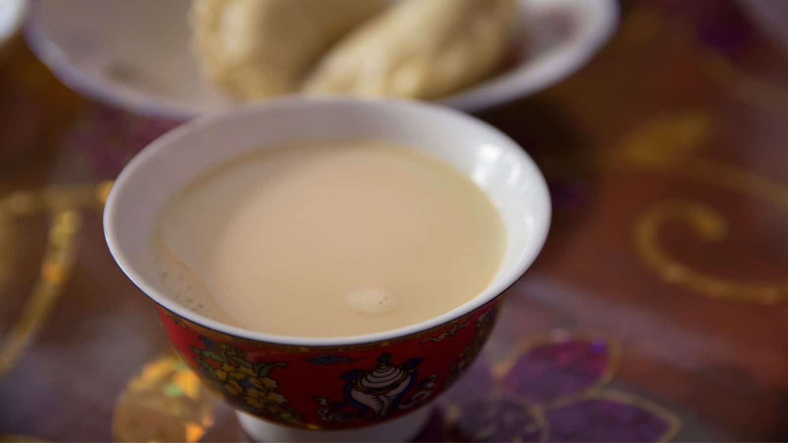 A cup of milky tea in a decorative red cup with floral patterns is on a table. In the background, there is a blurred image of a white, round pastry on a plate. The table has a colorful, floral-patterned tablecloth.