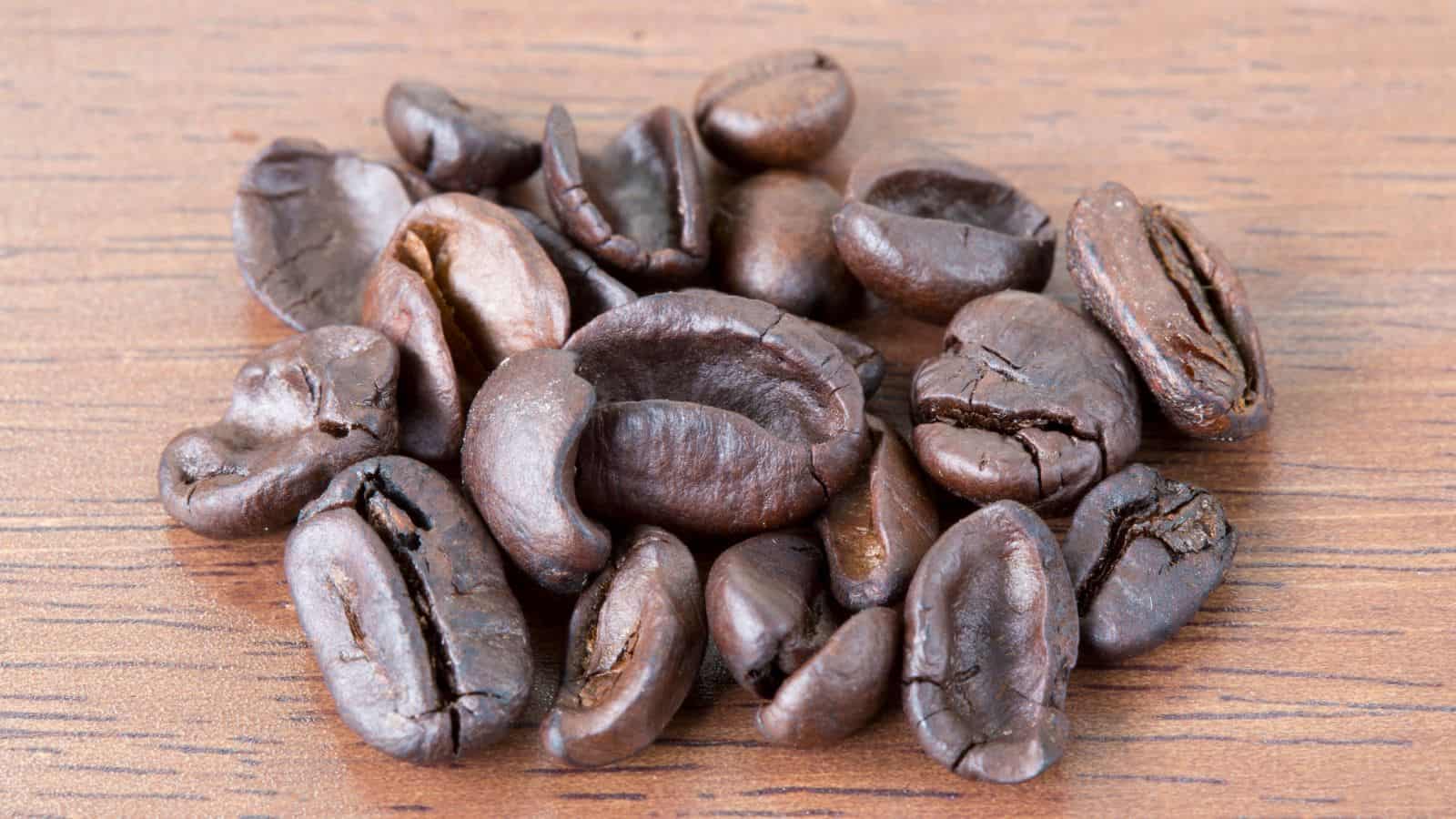 A close-up of roasted coffee beans scattered on a wooden surface. The beans are dark brown with a glossy sheen, showcasing their rich texture and natural cracks.