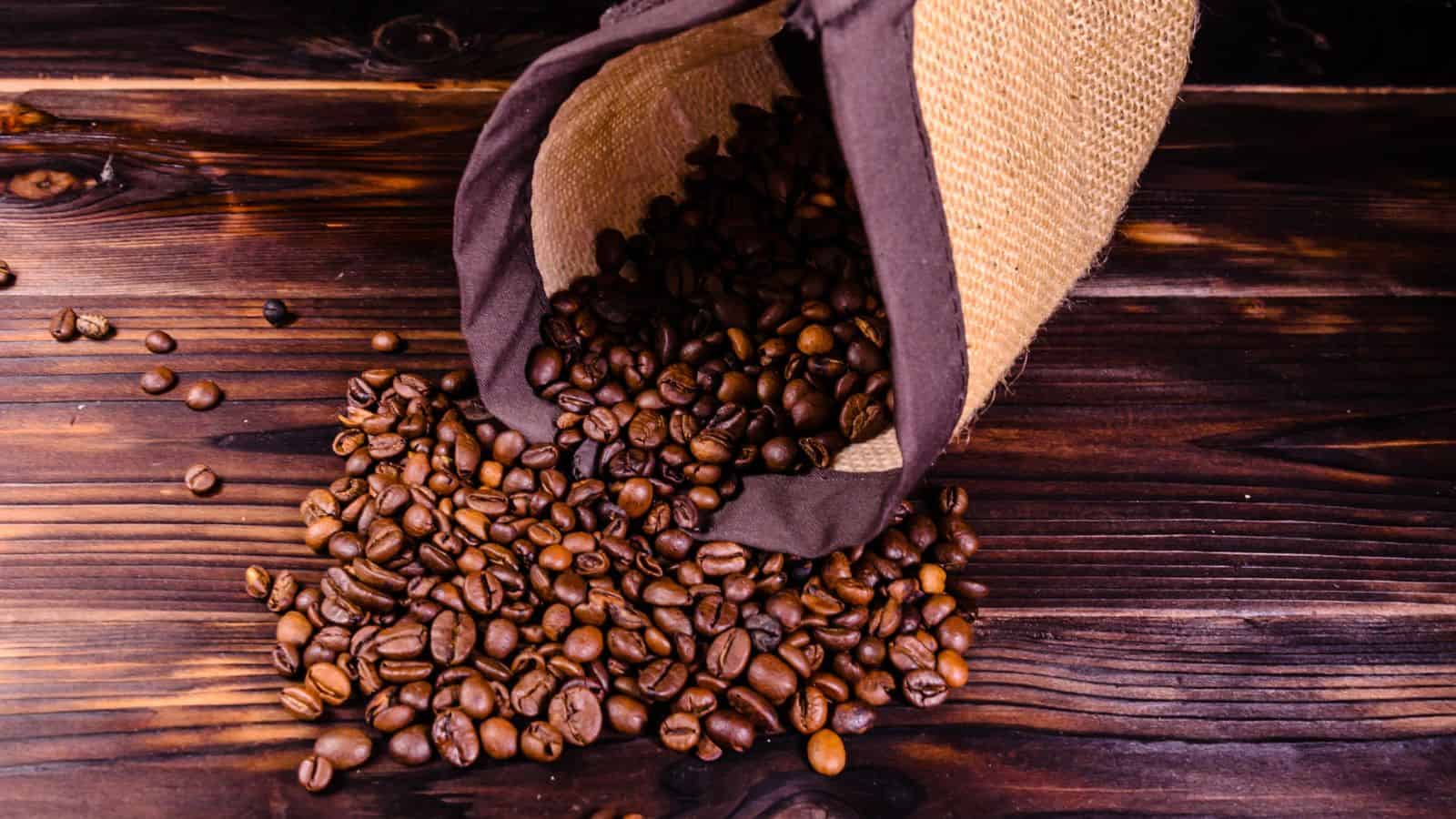 A burlap sack is tipped over on a wooden surface, spilling a pile of roasted coffee beans. The rich brown beans contrast with the darker wood background, creating a rustic and inviting appearance.