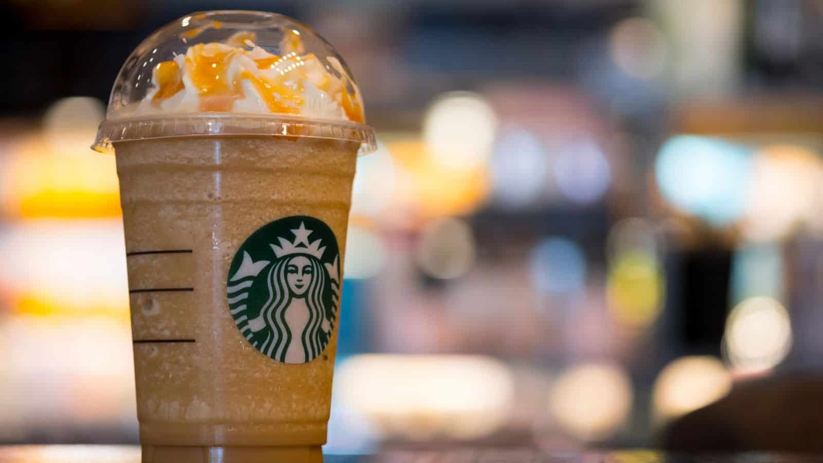 Close-up of a caramel Frappuccino in a clear cup with a domed lid and straw. The drink features whipped cream and caramel drizzle on top. The Starbucks logo is visible on the cup. The background is blurred, suggesting a caf&eacute; setting.