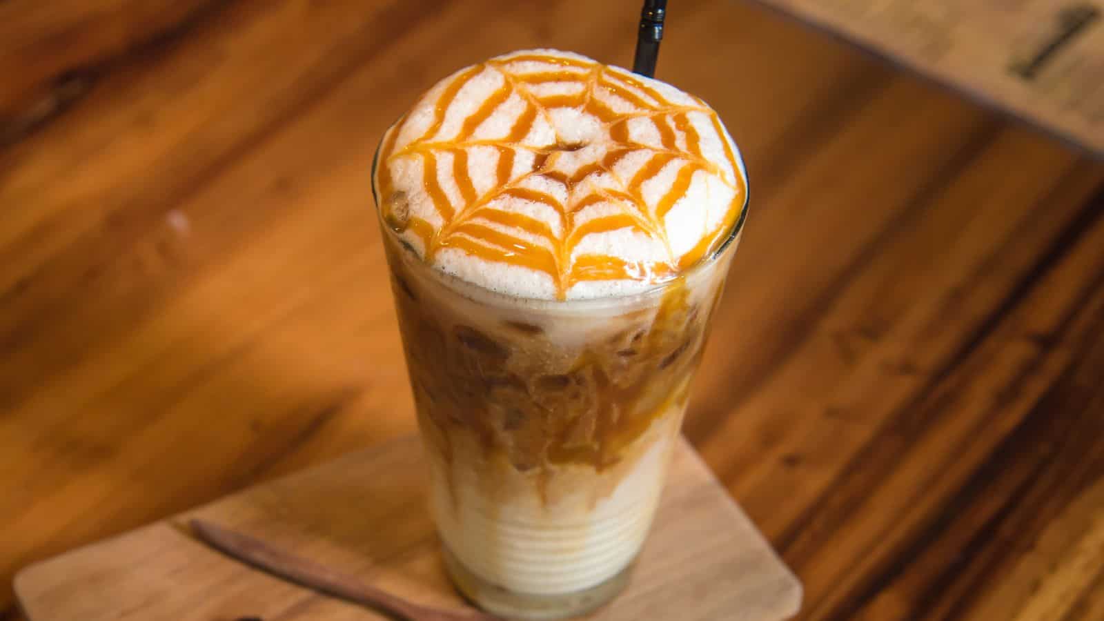 A tall glass of iced coffee topped with frothed milk and a decorative caramel drizzle sits on a wooden tray. A black straw is in the drink. The table is wooden with a warm finish.