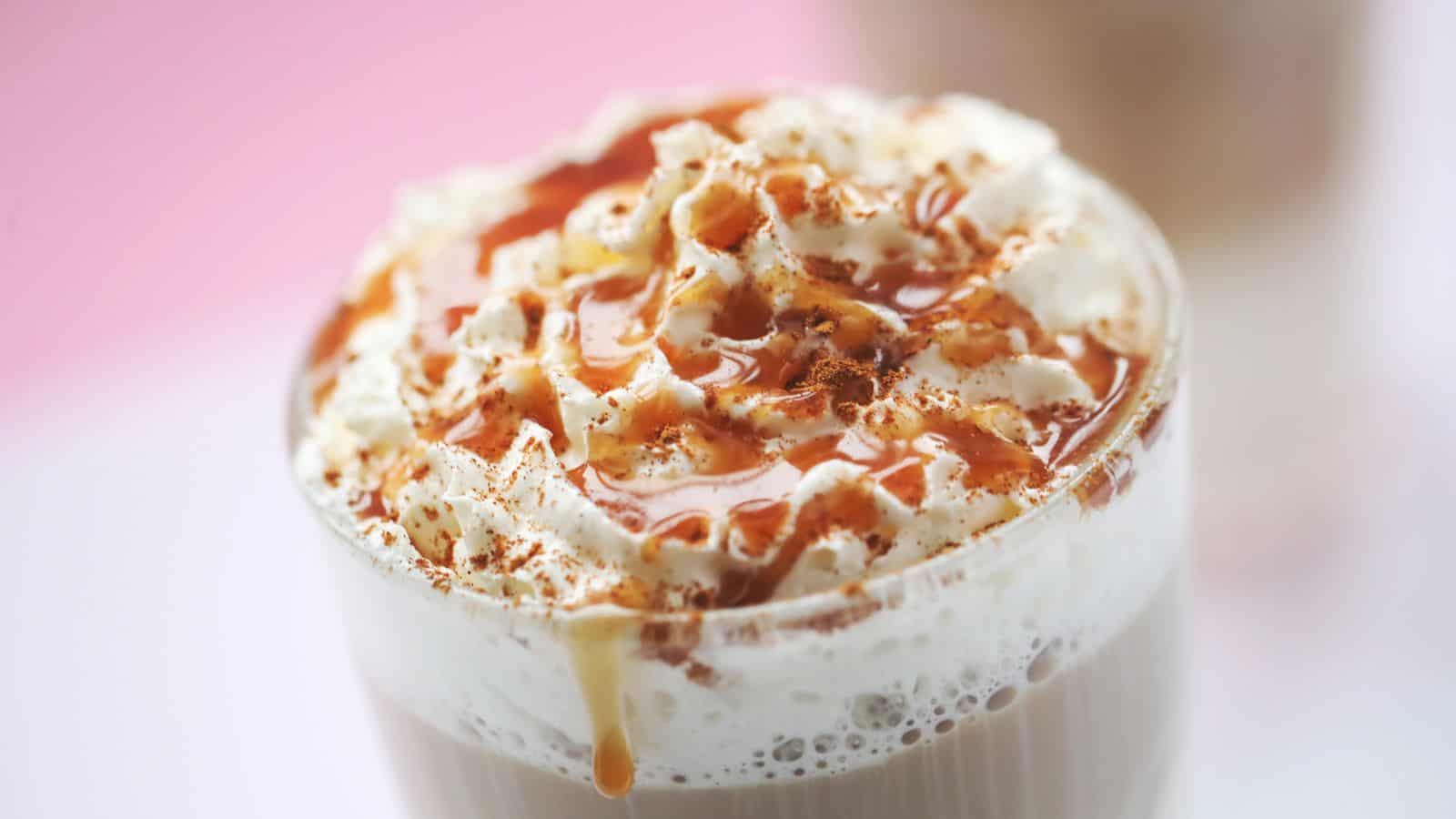 A close-up of a glass filled with a creamy beverage topped with whipped cream and drizzled with caramel sauce. The surface shows a light dusting of cinnamon. The background is softly blurred in pink tones.