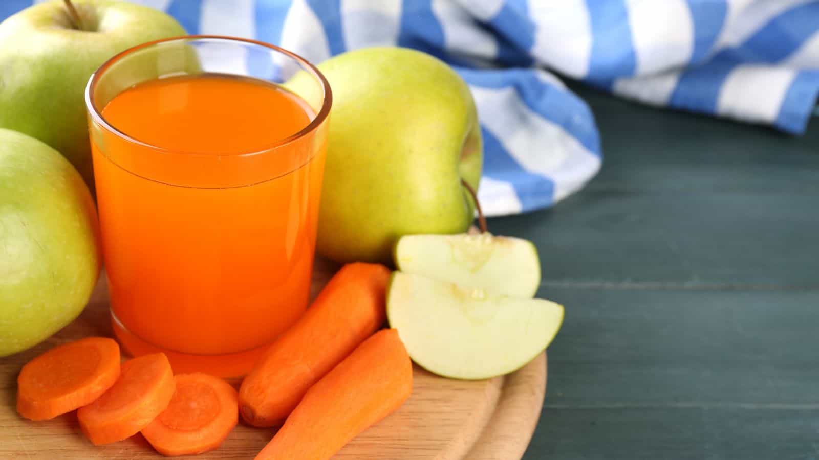 A glass of orange juice is placed on a wooden board surrounded by whole green apples, apple slices, and carrot slices. A blue and white striped cloth is visible in the background. The board is on a dark green surface.