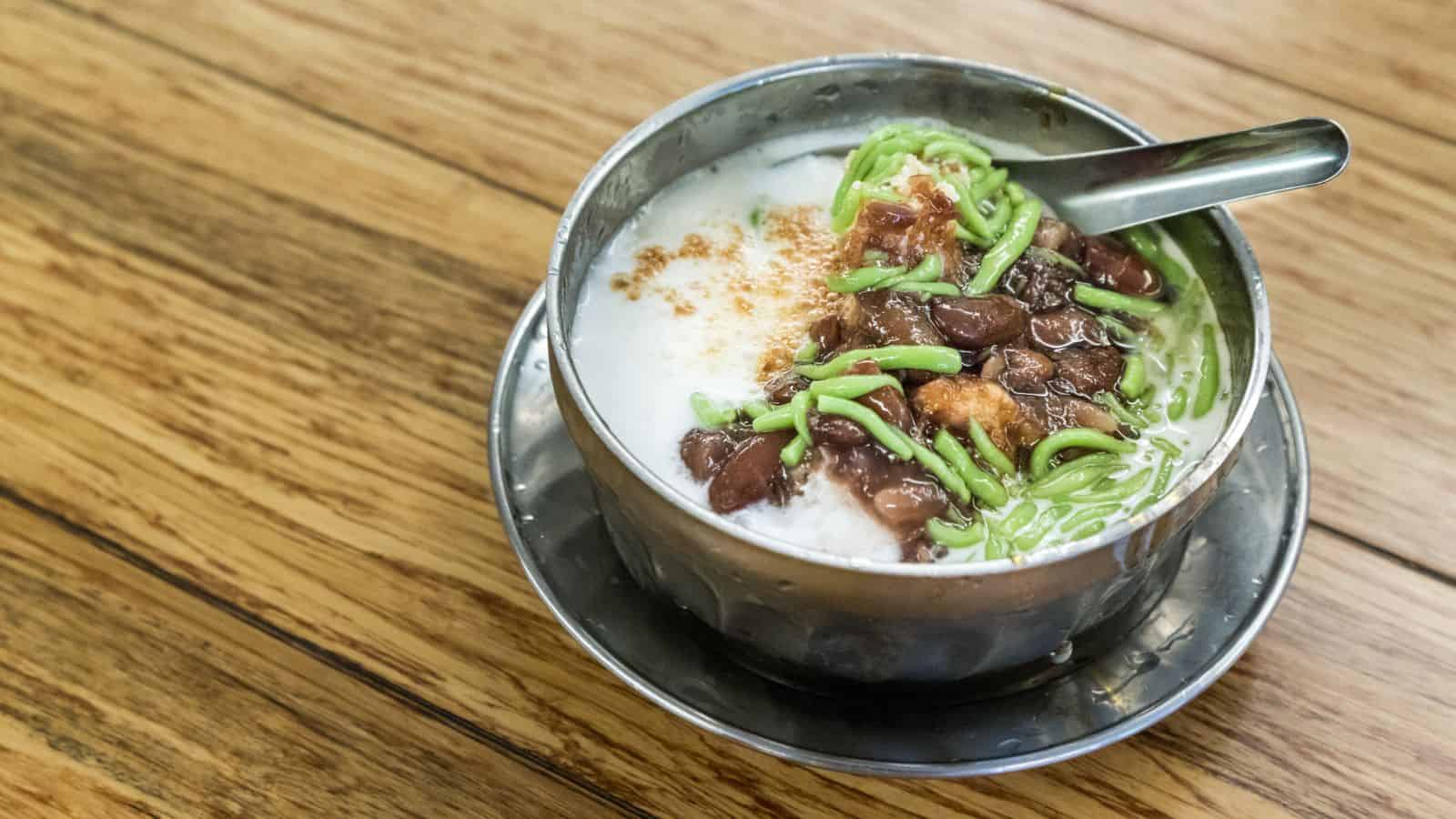 A metal bowl contains a dessert with green pandan noodles, red beans, and crushed ice topped with coconut milk. A spoon rests inside. The bowl is placed on a metal saucer on a wooden table.