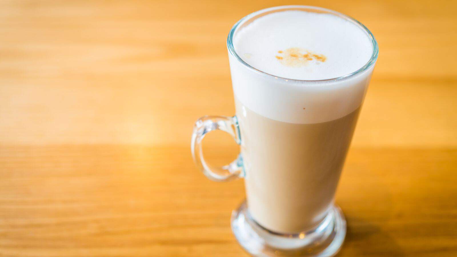 A clear glass mug filled with a frothy latte is placed on a wooden table. The latte has a layer of foam on top with a light brown spot in the center. The mug has a handle, and the table surface has a natural wood grain.