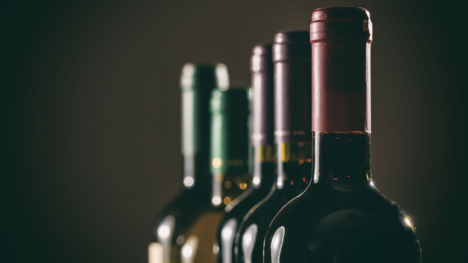 A row of four wine bottles with labels facing away. The focus is on the tops of the bottles, showing dark red, green, and dark purple foil capsule covers. The background is a dimly lit, blurred gradient, enhancing the silhouettes of the bottles.