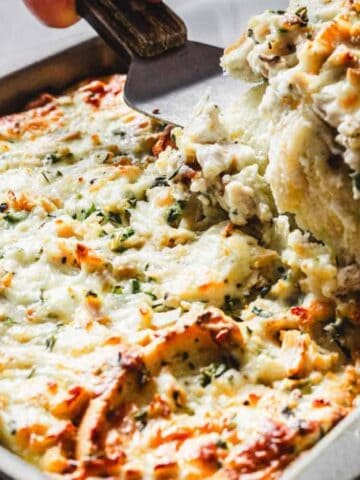 A person is using a spatula to serve a portion of a baked casserole from a rectangular metal dish. The casserole is topped with melted cheese and sprinkled with herbs.