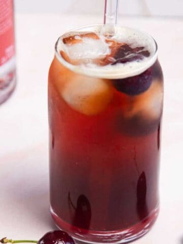 A tall, clear glass filled with a dark red drink, ice cubes, and garnished with cherries. A clear straw is inserted into the drink. Surrounding the glass are fresh cherries, with a bottle labeled "Italian Orange" in the background.