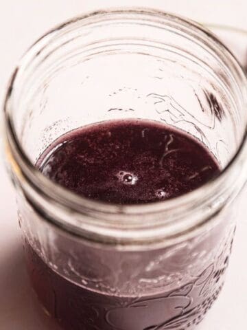 A glass jar filled with a dark red liquid sits on a light surface, viewed from above. Three fresh cherries with stems are placed nearby.