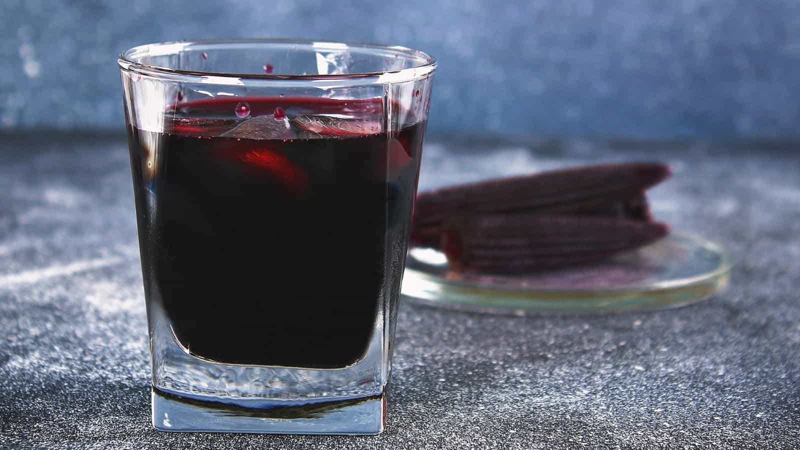 A glass of dark red juice is filled halfway, with ice cubes visible inside. In the background, there is a plate with several pieces of purple corn arranged on it. The surface is gray, and the background is softly blurred.
