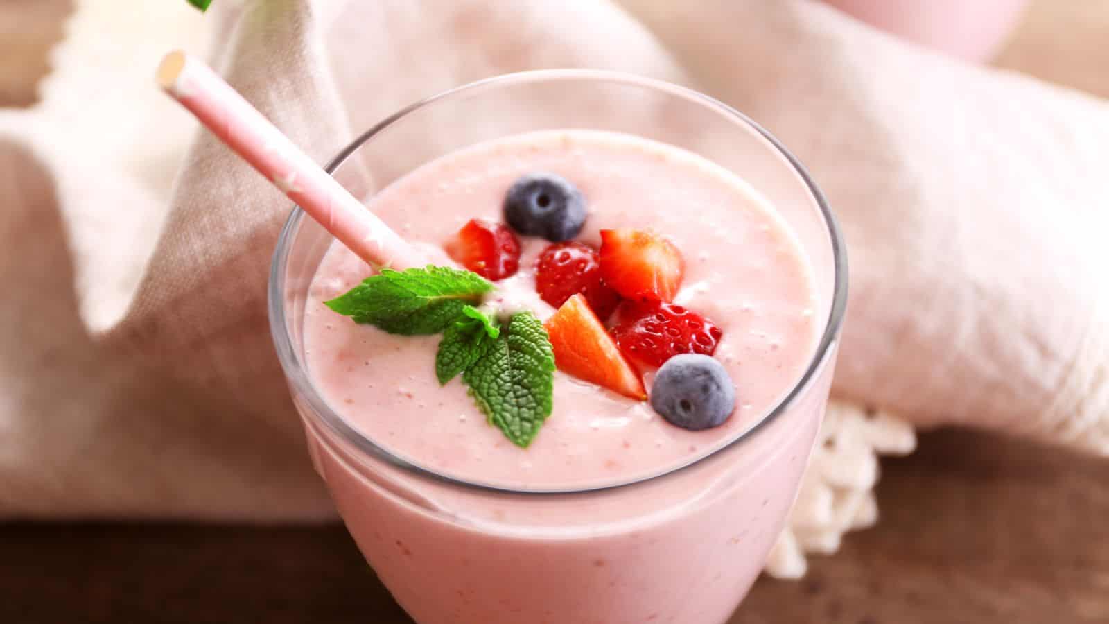 A glass of pink smoothie topped with fresh strawberries, blueberries, and mint leaves. A pink straw is placed in the glass. There is a cloth napkin in the background on a wooden surface.