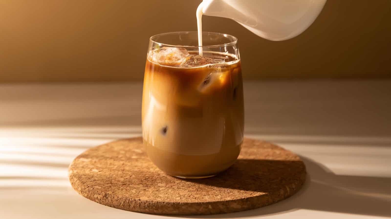 A glass of iced coffee is placed on a cork coaster. Milk is being poured into the glass, creating a swirling pattern as it mixes with the coffee. Sunlight casts soft shadows on the surface.