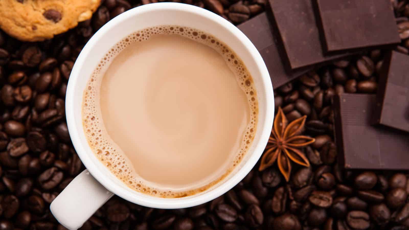 A white coffee cup filled with light brown coffee is surrounded by coffee beans, chocolate squares, a star anise, and a cookie, creating a cozy and warm atmosphere.