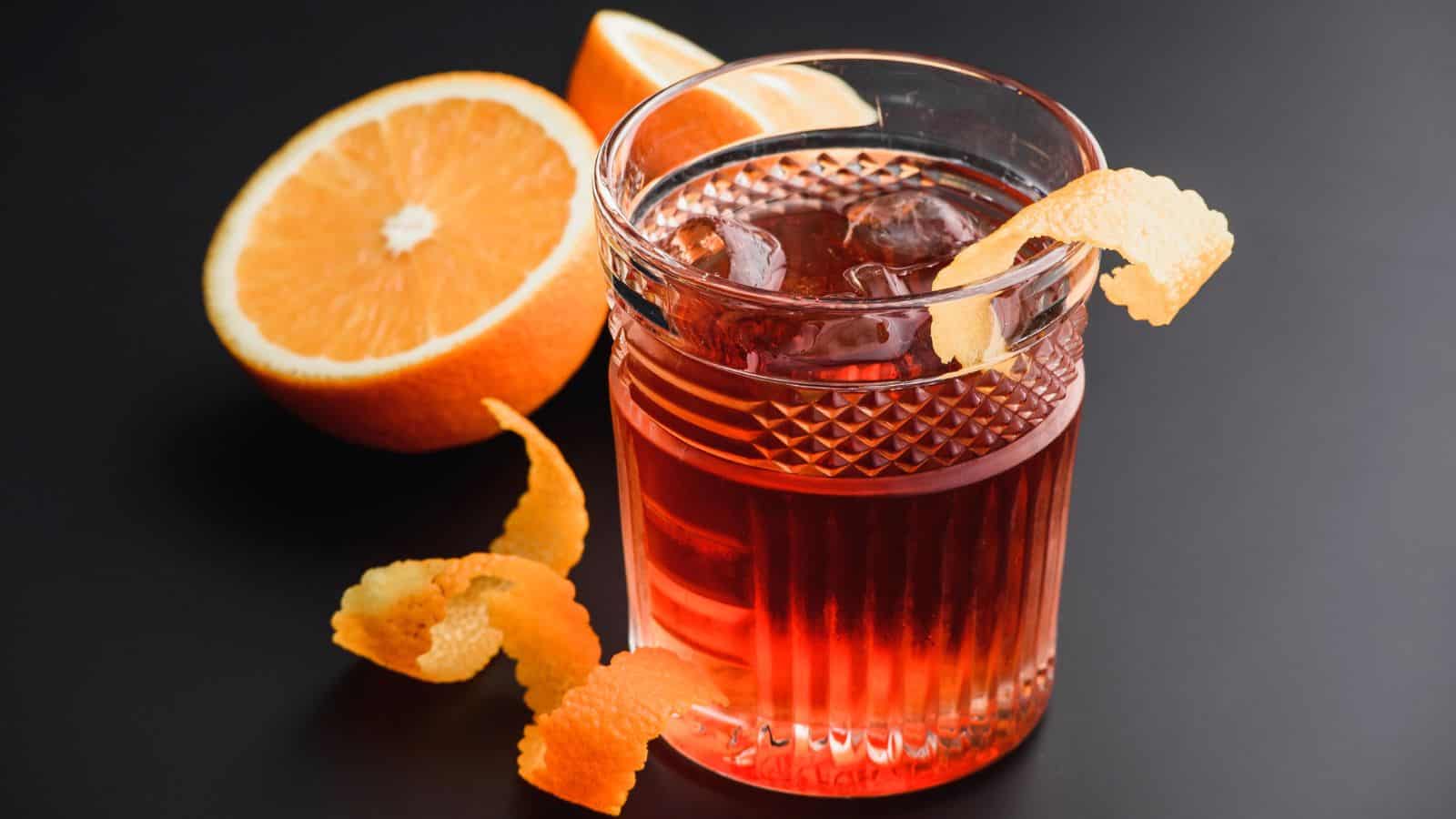 A glass filled with a reddish-orange cocktail and ice sits on a black surface. An orange peel garnish hangs over the rim. A halved orange and another orange peel are in the background.