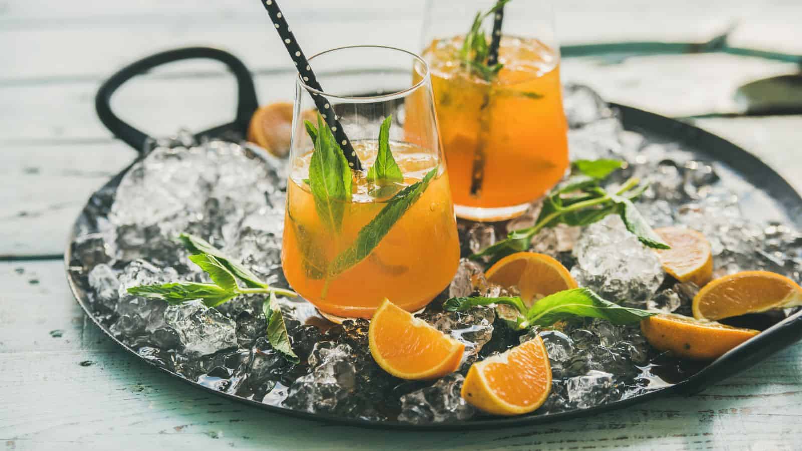Two glasses of orange cocktail garnished with mint leaves and dotted straws rest on a round tray filled with ice cubes. Sliced orange pieces and mint sprigs are scattered on the tray, set on a wooden surface.