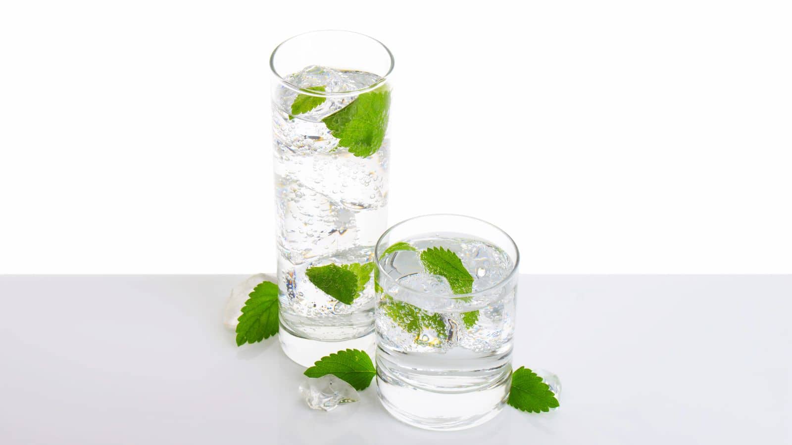 Two glasses filled with clear sparkling water, ice cubes, and mint leaves. One glass is tall and cylindrical, while the other is shorter and round. Ice cubes and mint leaves are scattered on the white surface around the glasses.