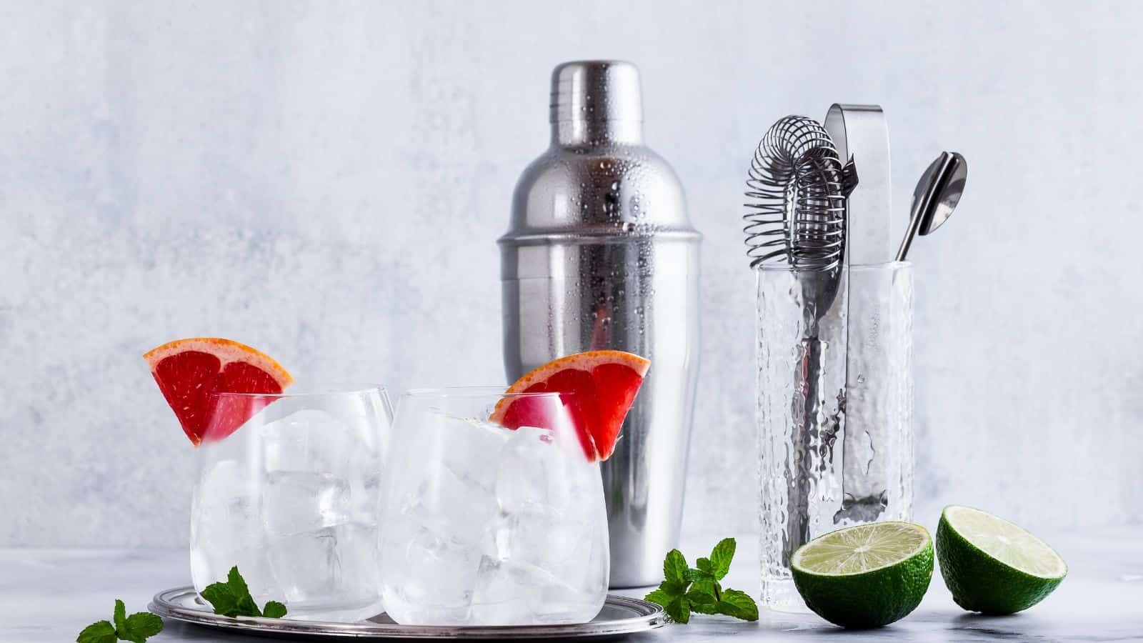 Two glasses filled with ice and garnished with red grapefruit slices sit on a tray. Beside them are a stainless steel cocktail shaker and a glass holding bar tools. A halved lime and mint leaves are on the table.
