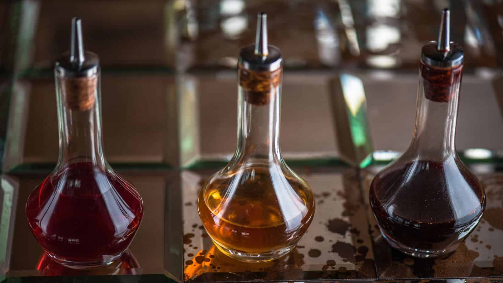 Three glass bottles with pour spouts, containing red, yellow, and dark brown liquids, are placed on a reflective square-tiled surface. The bottles have a similar shape with narrow necks and round bases. Some liquid spills are visible on the tiles.