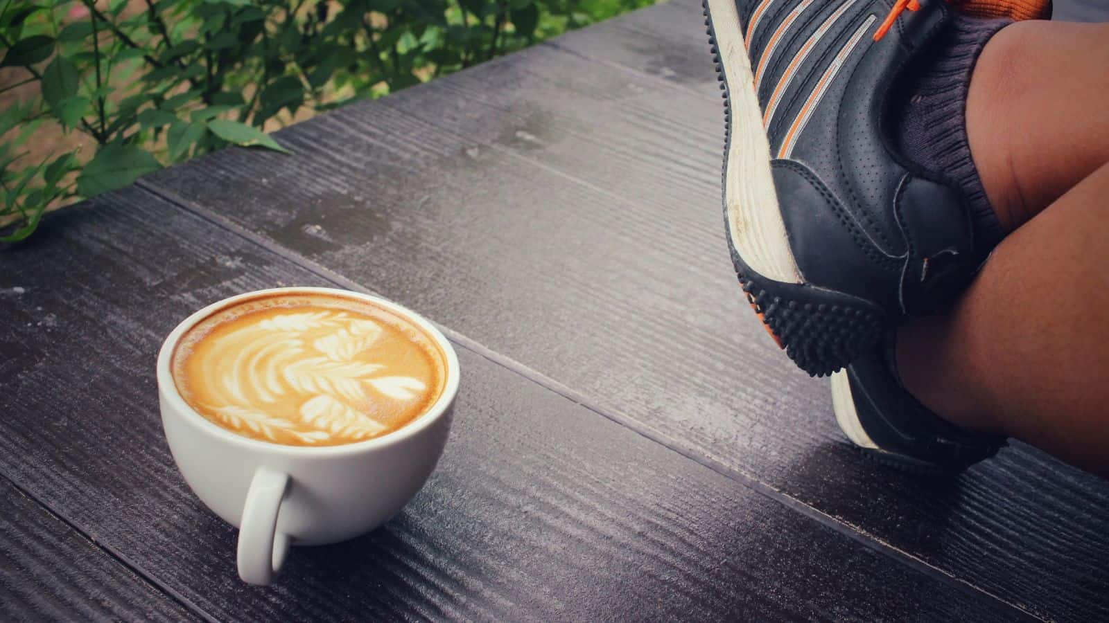A white cup filled with latte art sits on a dark wooden table. Next to the cup, a person's legs in black sneakers with white and orange stripes are crossed and resting on the table. Green leaves are in the background.