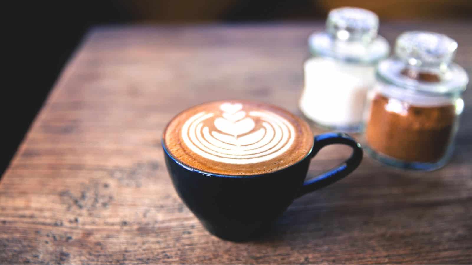 A black ceramic mug filled with a latte, featuring intricate latte art on top, sits on a wooden table. Two glass jars containing sugar and cocoa powder are in the background.