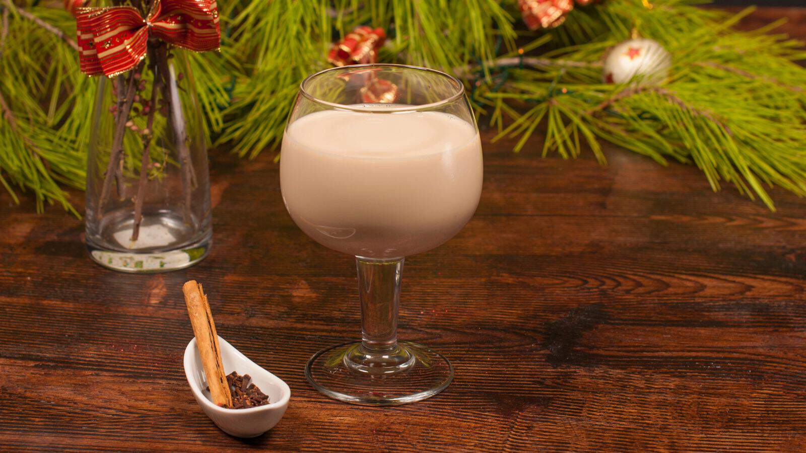 A glass of creamy beverage on a wooden table. Next to it is a small dish containing cinnamon sticks and cloves. In the background, there are green pine branches and a vase with a red ribbon.