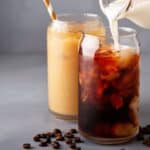 Two glasses of iced coffee on a gray surface. One glass has milk being poured into it, creating a swirling effect. The other glass has coffee with a straw. Coffee beans are scattered on the surface nearby.