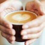 A person holds a brown cup filled with coffee, featuring a heart-shaped latte art on the surface. The focus is on the cup and hands, with a blurred background.
