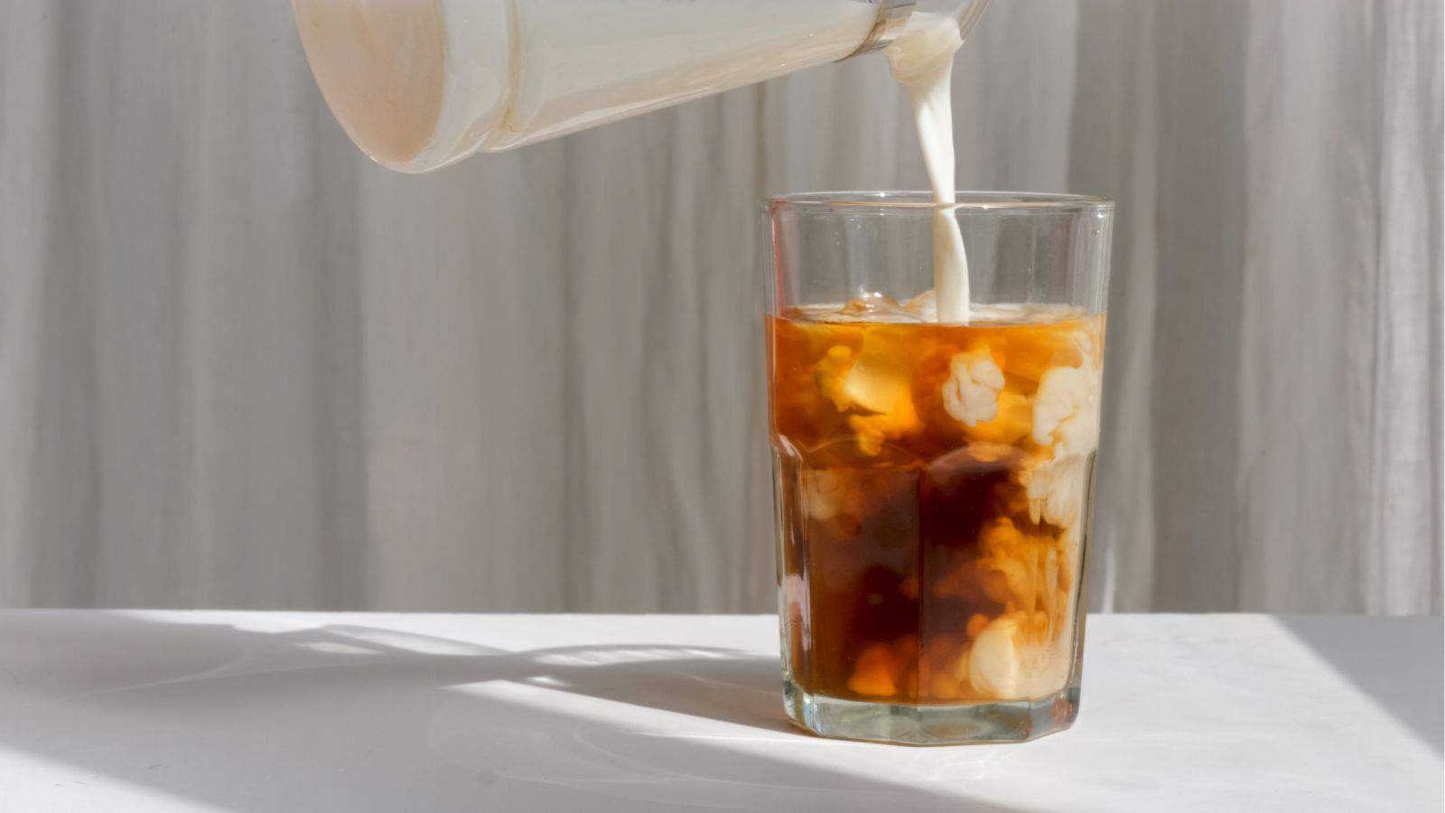 Milk is being poured into a glass of iced coffee on a white surface. The glass is filled with ice cubes and the milk is creating a swirling effect in the coffee. A sheer curtain is in the background.