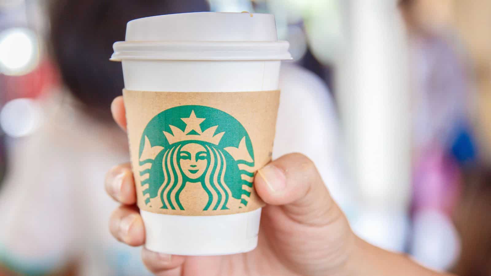 A hand holding a white paper coffee cup with a brown sleeve featuring the green Starbucks logo. The background is blurred, suggesting an indoor setting.