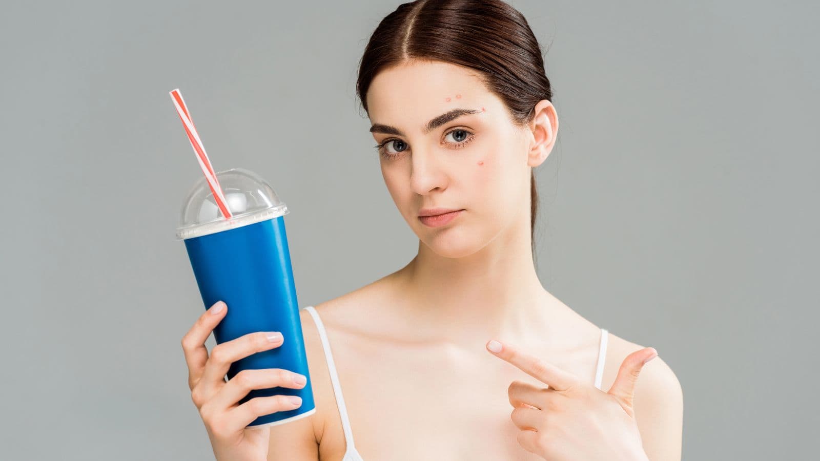 A woman with dark hair is holding a blue cup with a red and white striped straw and a clear dome lid. She is pointing at a blemish on her forehead. The background is plain and gray.