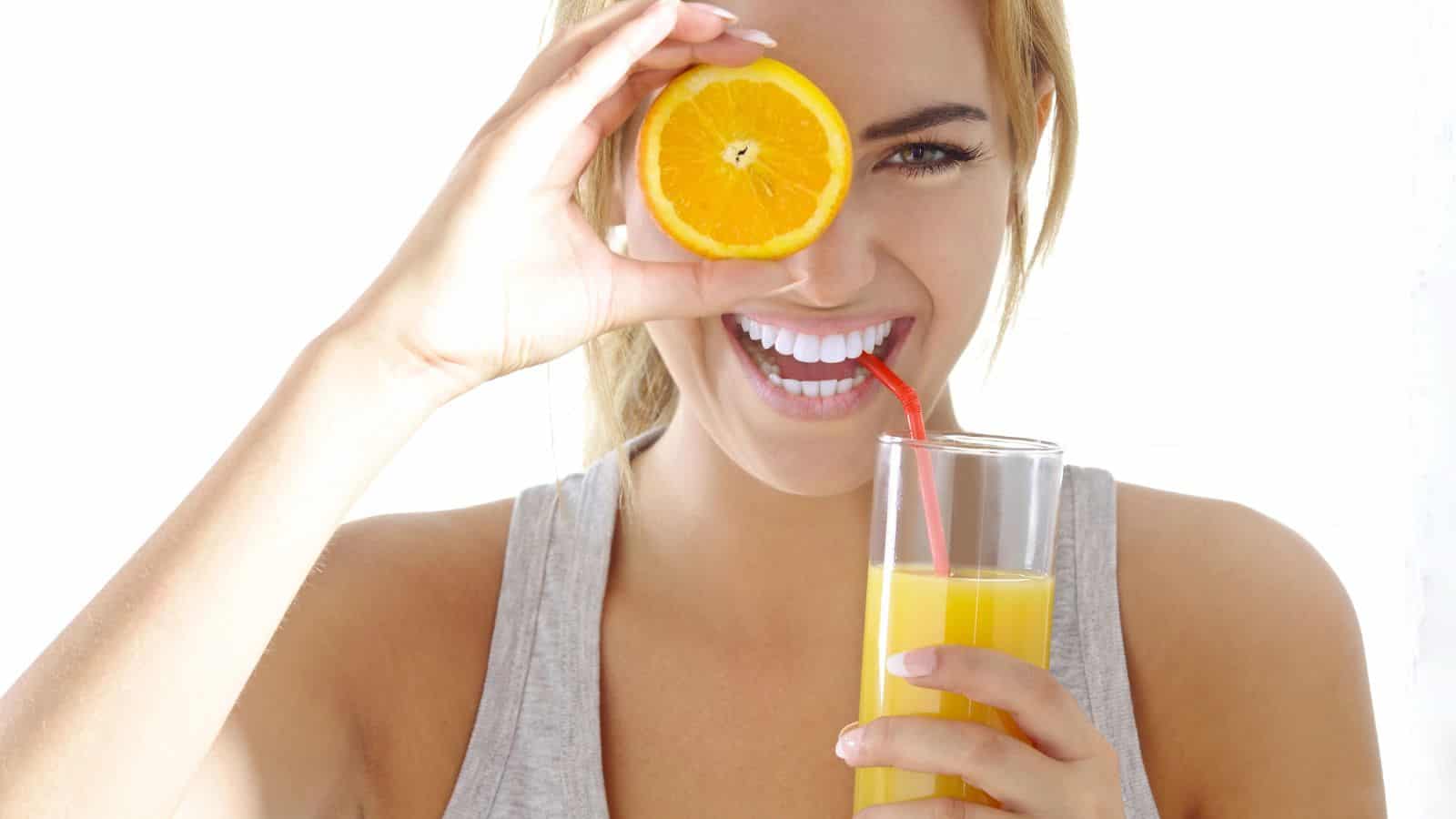 A woman holding a glass of orange juice with a red straw smiles while holding half an orange in front of one eye. She is wearing a gray tank top against a bright white background.