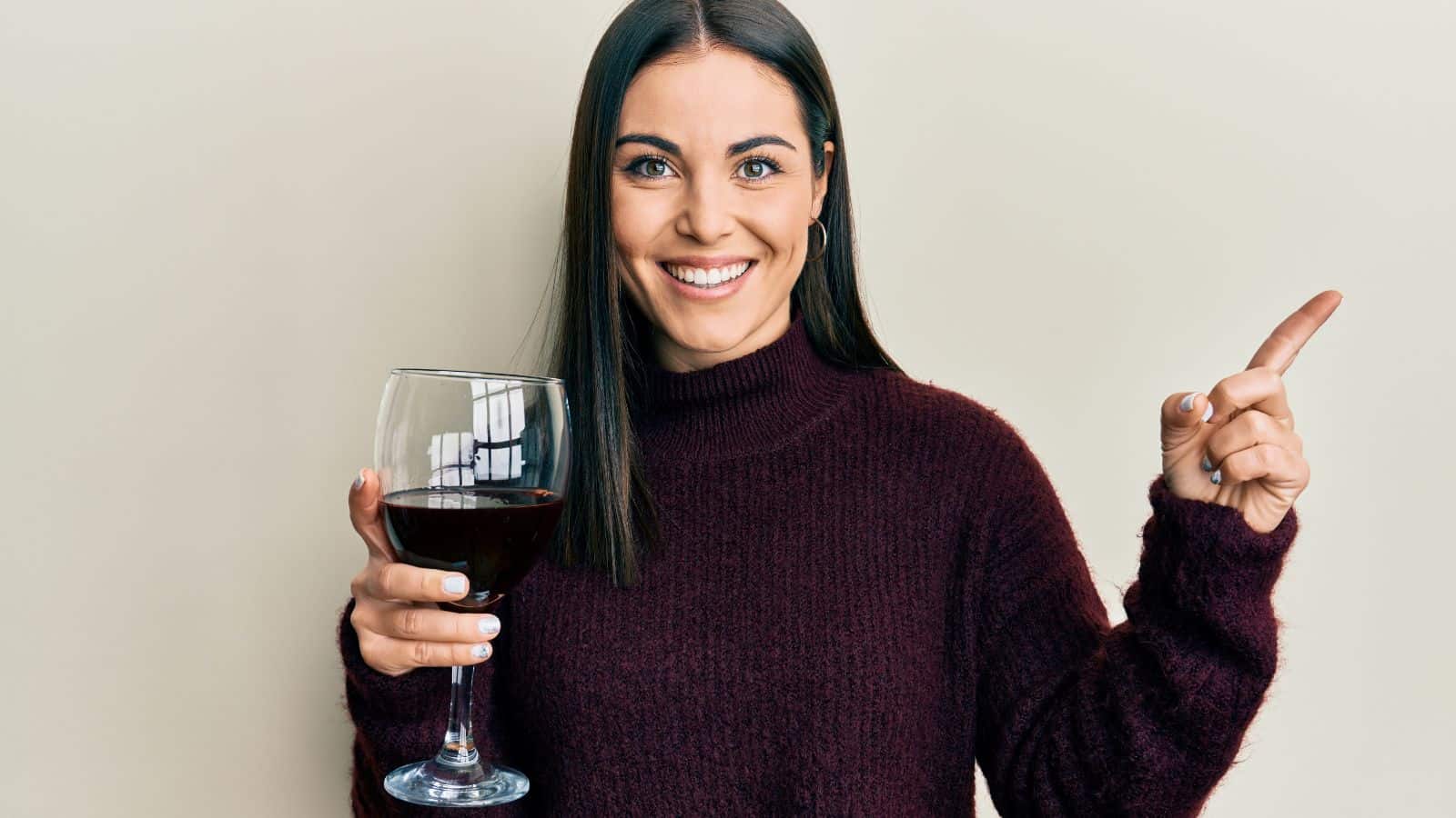 A person with long brown hair, wearing a dark maroon sweater, is holding a glass of red wine in their right hand and pointing upwards with their left hand. They are smiling and standing against a light beige background.