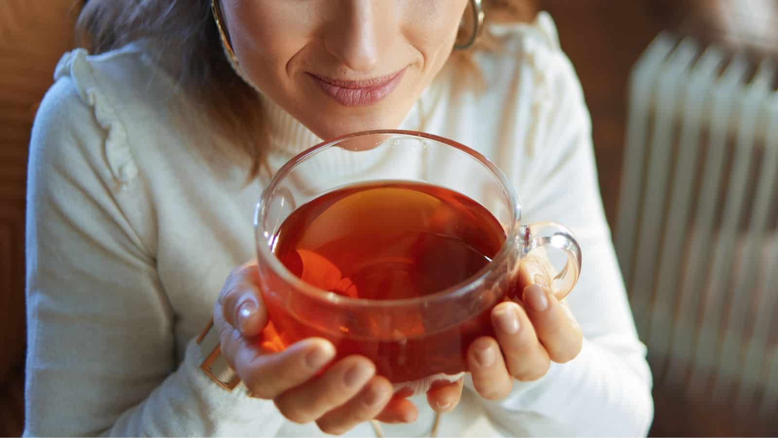 A person in a white sweater is holding a clear glass cup filled with tea. The cup is held close to their face, and they appear to be enjoying the aroma. The background is softly blurred.