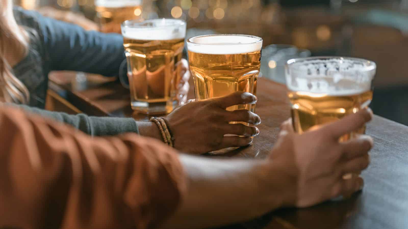 Three people hold glasses of beer at a wooden bar counter. The glasses are filled with amber-colored beer, and the foam is visible on top. The atmosphere suggests a casual, social setting.