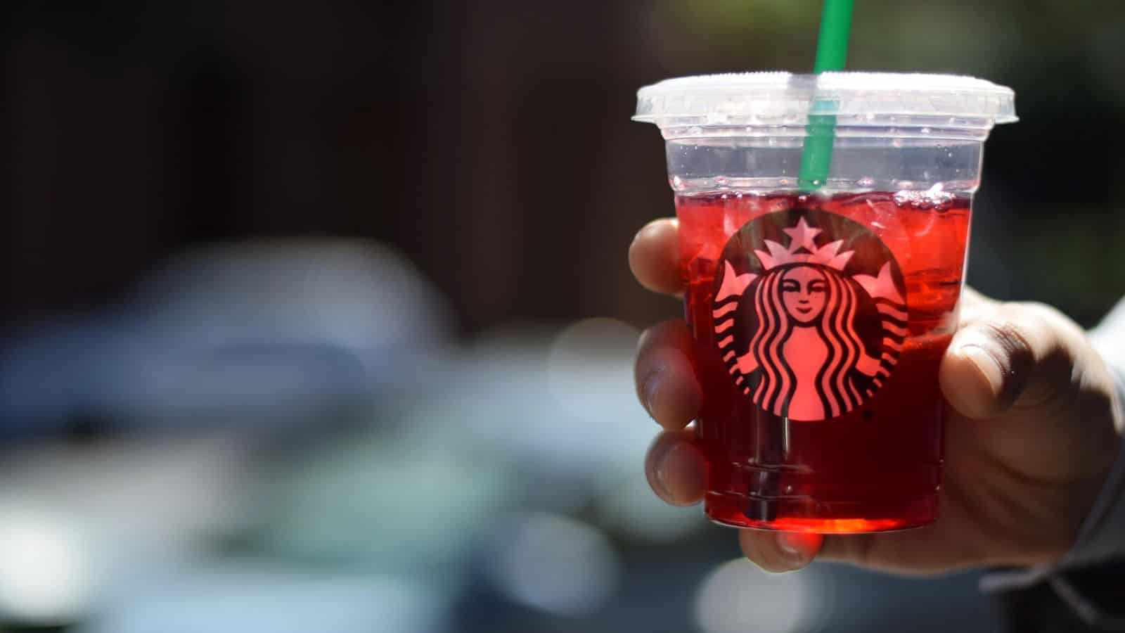 A hand holding a clear plastic cup with a green straw, filled with a red iced beverage. The cup features the Starbucks logo. In the blurred background, there are hints of street elements and parked cars.
