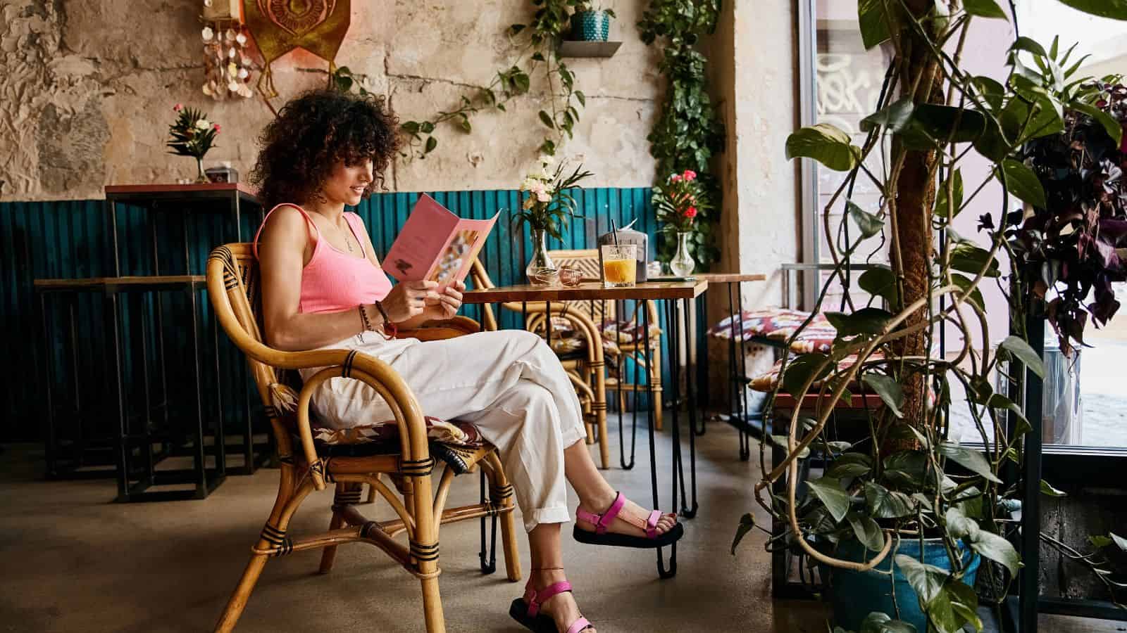 A person with curly hair sits in a wicker chair at a cafe, reading a book. They are wearing a pink top, white pants, and pink sandals. The cafe has wooden tables, potted plants, and a stone wall with a decorative mask.