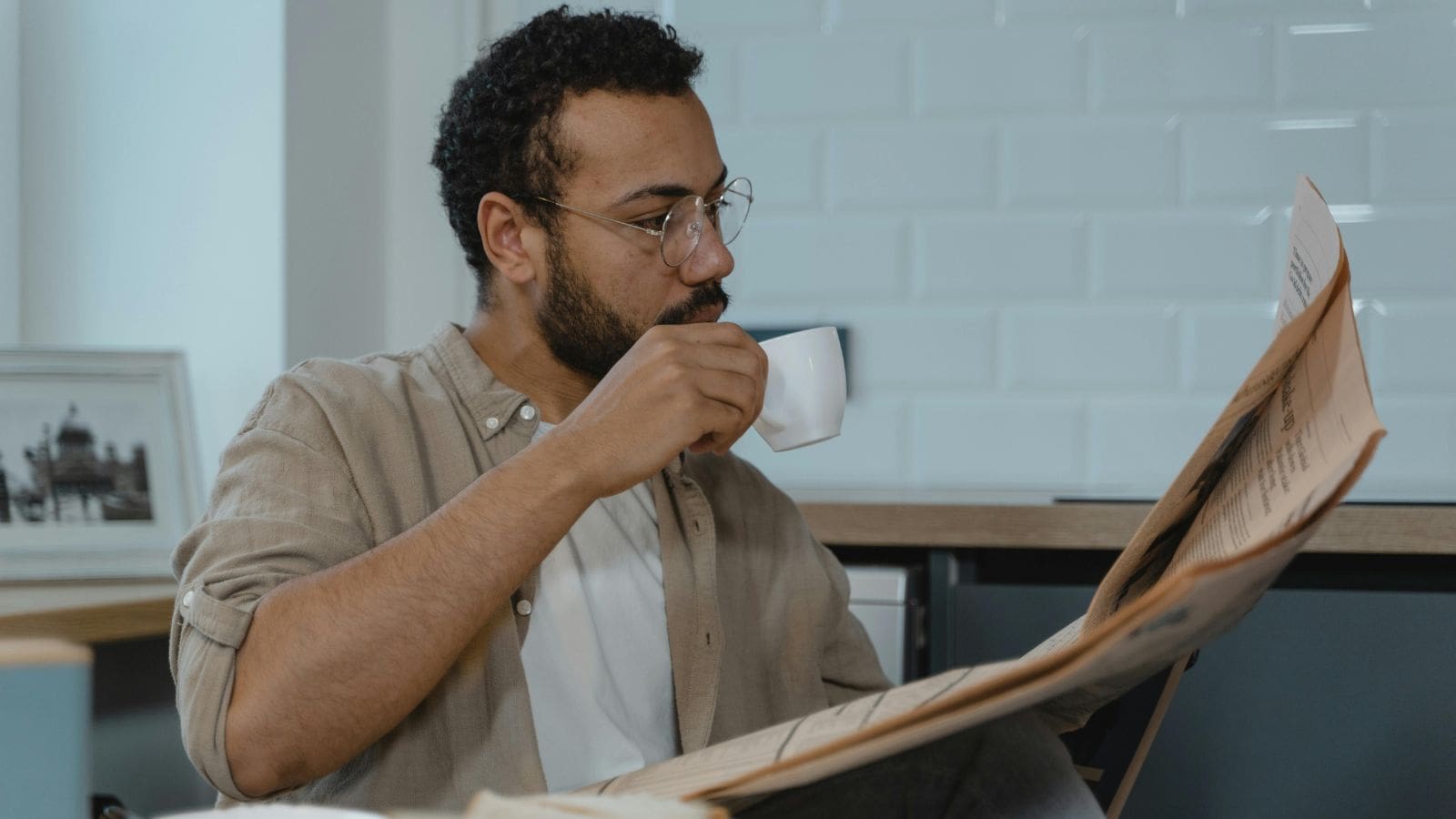 A man with glasses is reading a newspaper while sitting indoors. He is holding a small cup and taking a sip. There is a photo frame in the background, and he wears a beige shirt over a white t-shirt.