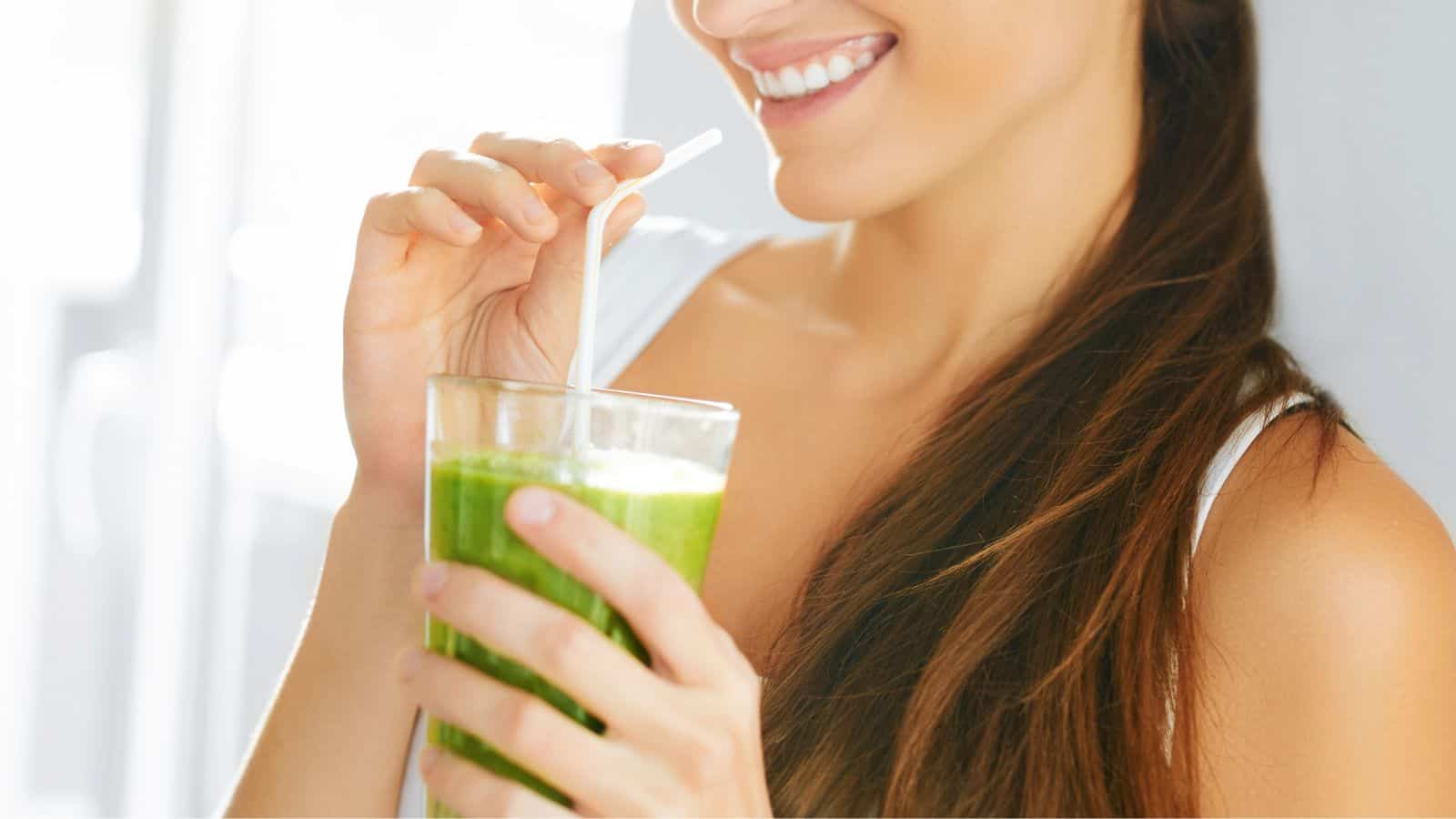 A person is smiling and holding a glass filled with a green smoothie, sipping through a straw. The focus is on the glass and the person's hands, with a clear view of the smoothie. The background is softly lit.