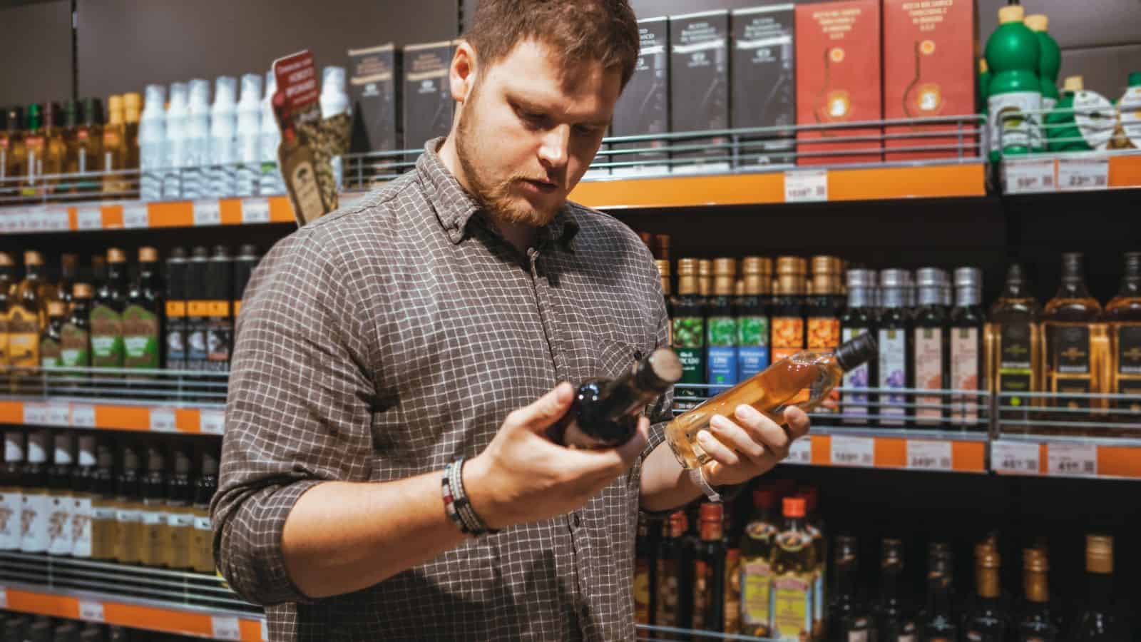 A man with a beard is in a store aisle holding a bottle in each hand, inspecting them. Shelves behind him are stocked with various alcoholic beverages, including whiskey, vodka, and wine. He is wearing a checked shirt.