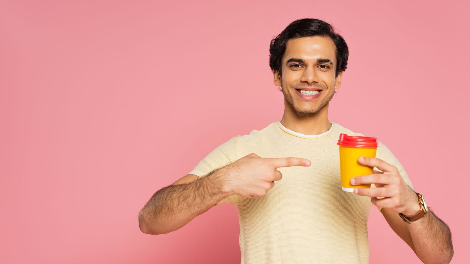 A smiling person in a yellow shirt stands against a pink background, holding a yellow cup with a red lid in one hand and pointing to it with the other.