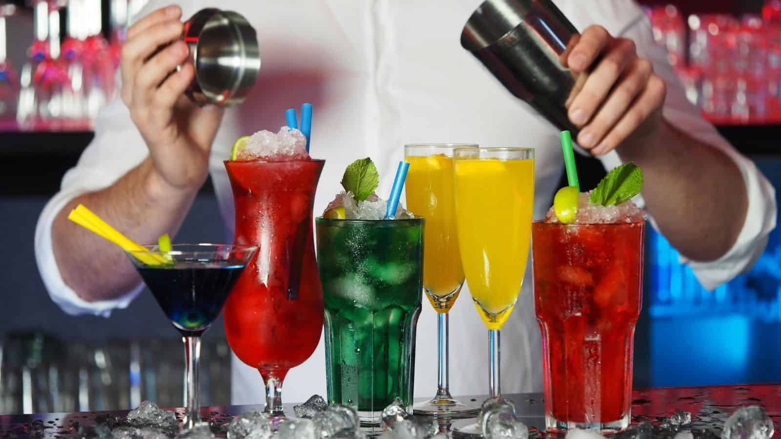 A bartender prepares drinks behind a bar counter. In front, there are various colorful cocktails: one blue with a lemon slice, two red with straws, one green with mint leaves, and two yellow with straws. Ice cubes are scattered on the counter.